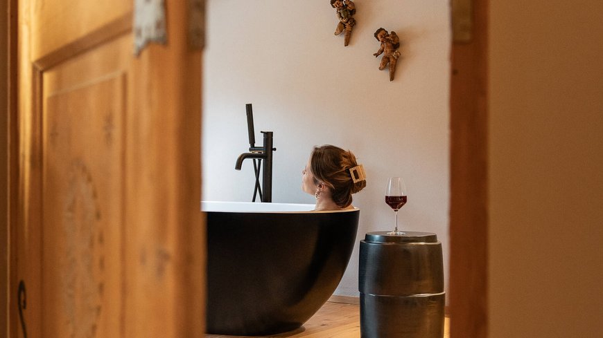 Schwarzer Adler Sterzing: A historic icon. Reinvented. Frau entspannt in schwarzer Badewanne, mit Weinglas auf Hocker, gesehen durch Tür