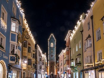 Schwarzer Adler Sterzing: A historic icon. Reinvented. Beleuchtete Fußgängerzone mit historischen bunten Häusern und Turmuhr bei Nacht