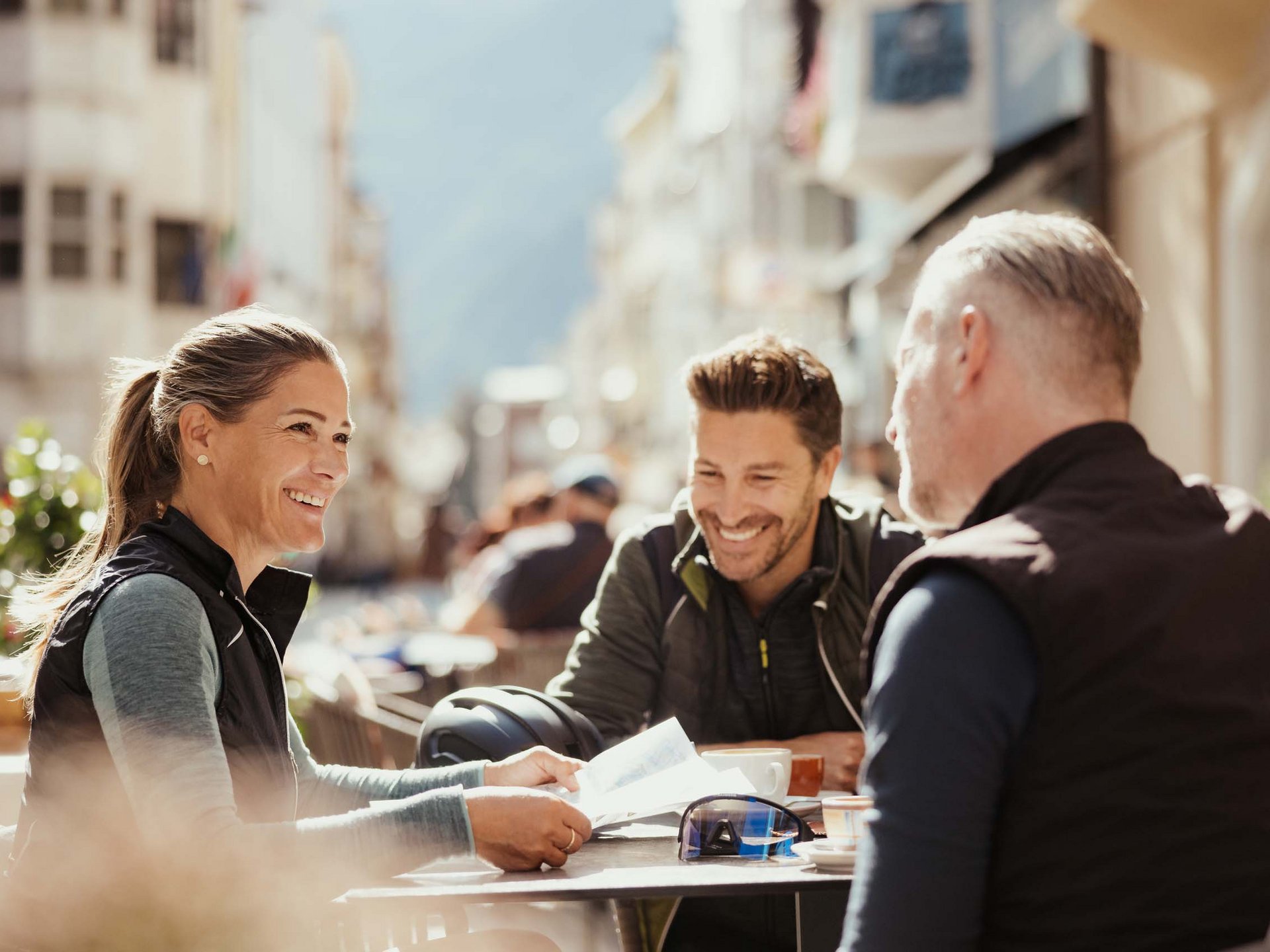 Cooles Rennradhotel in Südtirol Drei lächelnde Menschen sitzen im Freien an einem Tisch mit Getränken.