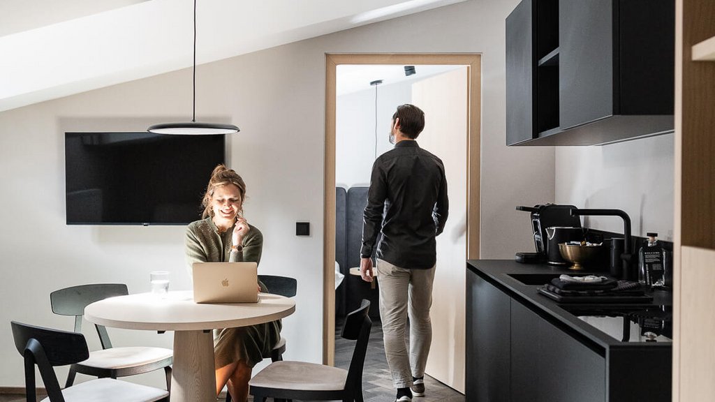 Schwarzer Adler Sterzing: A historic icon. Reinvented. Frau arbeitet mit Laptop am Tisch, Mann geht durch Zimmertür in moderner Wohnung