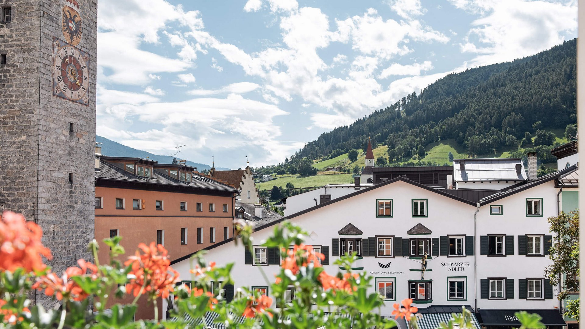 The perfect location for your apartment in Sterzing Old town with clock tower, flowers, and mountains in the background on a sunny day