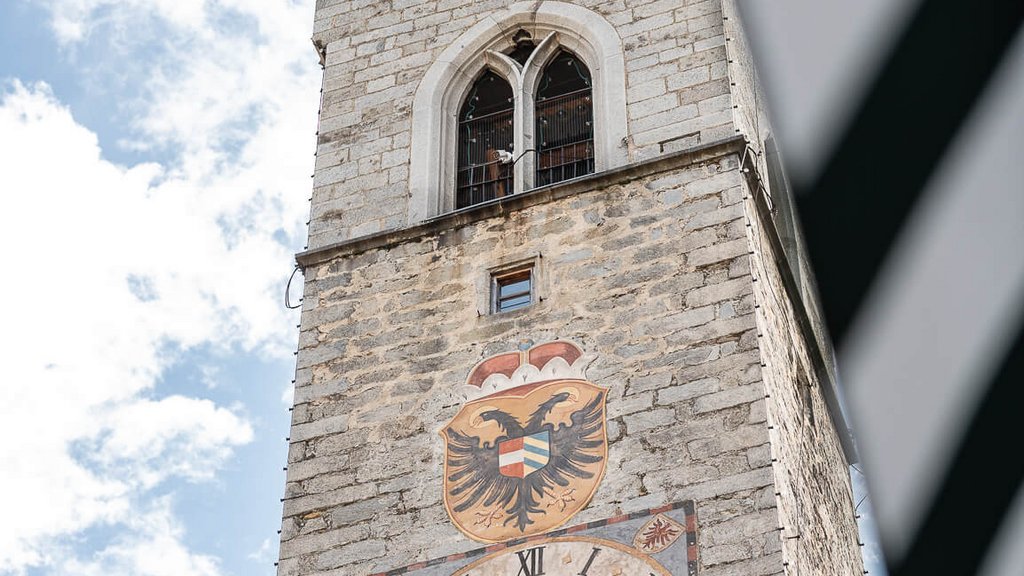 The perfect location for your apartment in Sterzing Historic stone clock tower with coat of arms under blue sky
