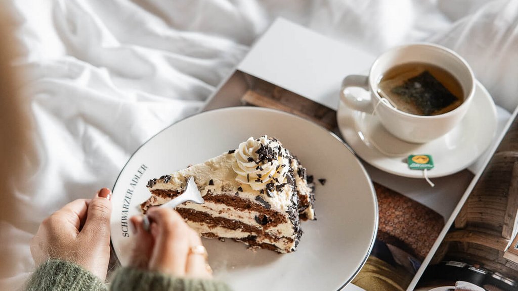 Schwarzer Adler Sterzing: A historic icon. Reinvented. Hand hält Teller mit Stück Kuchen neben Tasse Tee auf Bett mit Magazin