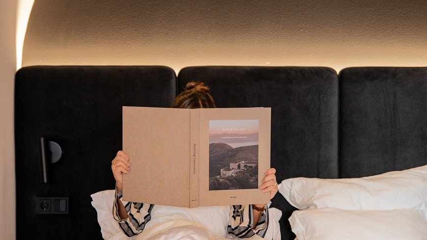 Schwarzer Adler Sterzing: A historic icon. Reinvented. Person liest Buch im Bett mit schwarzem Kopfteil und warmem Lichtbogen