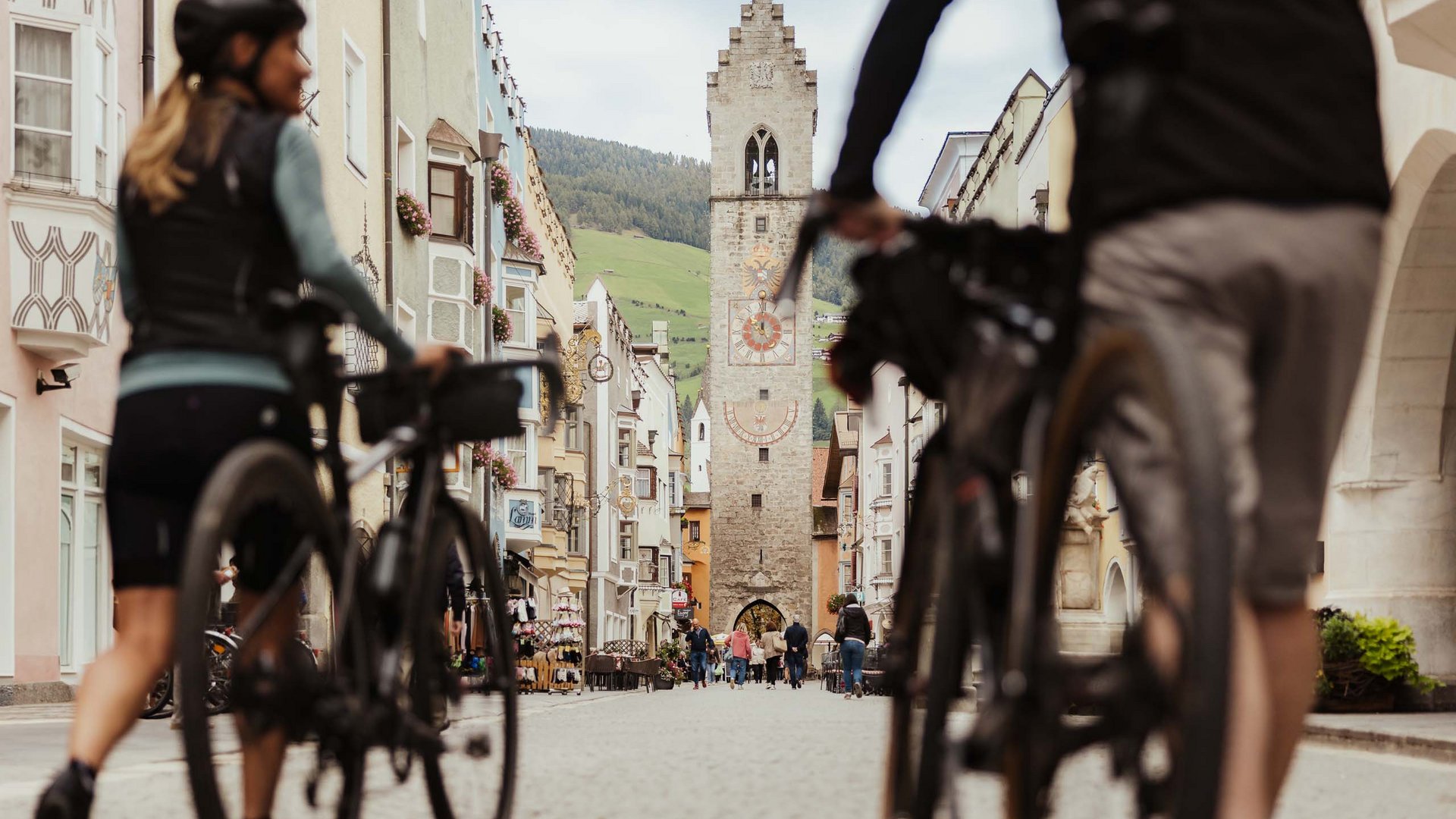 Cooles Rennradhotel in Südtirol Zwei Radfahrer gehen durch eine Altstadtstraße mit historischem Turm im Hintergrund